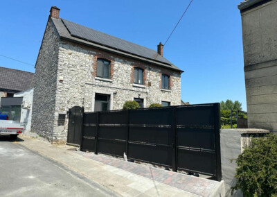 Aménagement extérieur d'une maison en pierre avec toiture équipée de panneaux solaires et portail noir moderne